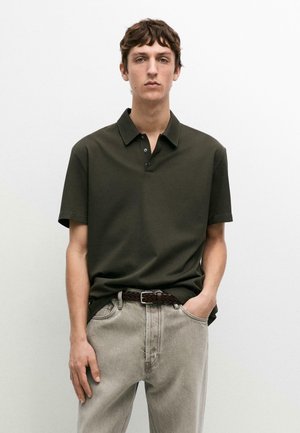 Giovane uomo con capelli corti castani che indossa una polo a maniche corte verde scuro e jeans grigi chiari, in piedi con una mano in tasca.