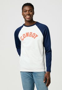 Weißes Langarm-T-Shirt mit navyfarbenen Raglanärmeln. "COWBOY" in Koralle quer über die Brust gedruckt. Gerade Passform, aus glattem Stoff.