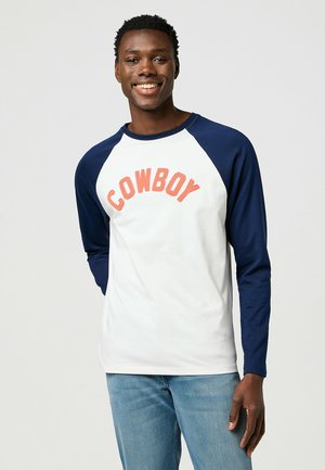 Weißes Langarm-T-Shirt mit navyfarbenen Raglanärmeln. "COWBOY" in Koralle quer über die Brust gedruckt. Gerade Passform, aus glattem Stoff.