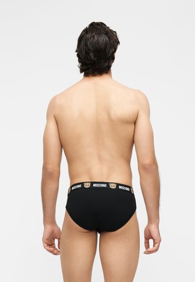 Άνδρας που φαίνεται από πίσω φοράει μαύρα εσώρουχα με το λογότυπο Moschino και εικονίδια αρκουδάκια στη μέση, σε απλό γκρι φόντο.