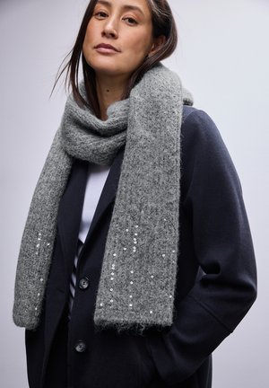 Écharpe en tricot gris avec une finition texturée, ornée de sequins le long du bord, portée par-dessus un manteau sombre à fermeture à bouton.