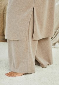 Beige dvoudílná souprava loungewear obsahující dlouhý top s bočními rozparky a široké kalhoty. Měkká tkanina, uvolněný vzor a jednoduchý design.