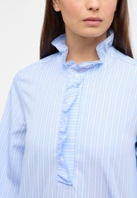 Helles blaufarbenes gestreiftes Blusenshirt mit Rüschensaum und Knopfleiste, das vertikale Nadelstreifen in einer weichen Textur aufweist.