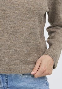 Beige kötött pulóver puha textúrával, hosszú ujjaival és kerek nyakkivágással, farmereken viselve.