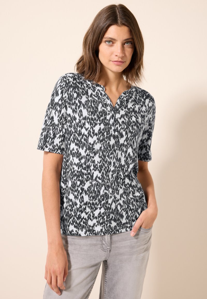 Camiseta de manga corta con un patrón abstracto en blanco y negro, que presenta un diseño sin cuello y una tela suave. Combinada con jeans gris claro.