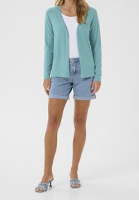 Kobieta ubrana w biały top, otwarty, morski kardigan, jasnoniebieskie, podwinięte szorty jeansowe oraz sandały na obcasie z denimu na białym tle.