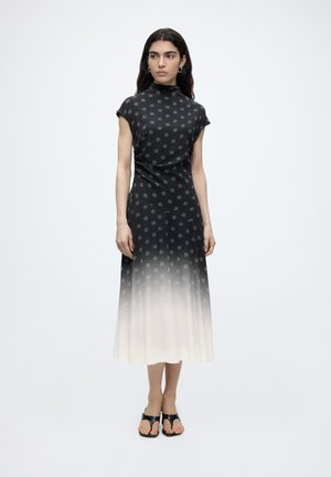 Femme portant une robe dégradée noir et blanc avec de petits motifs clairs, debout, les bras détendus, portant des sandales noires à bouts ouverts.