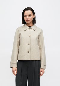 Beige jakke i ullblanding med krage, lukking med fem knapper og sidelommer. Har rett snitt og oppbrettede ermer, kombinert med mørke bukser.