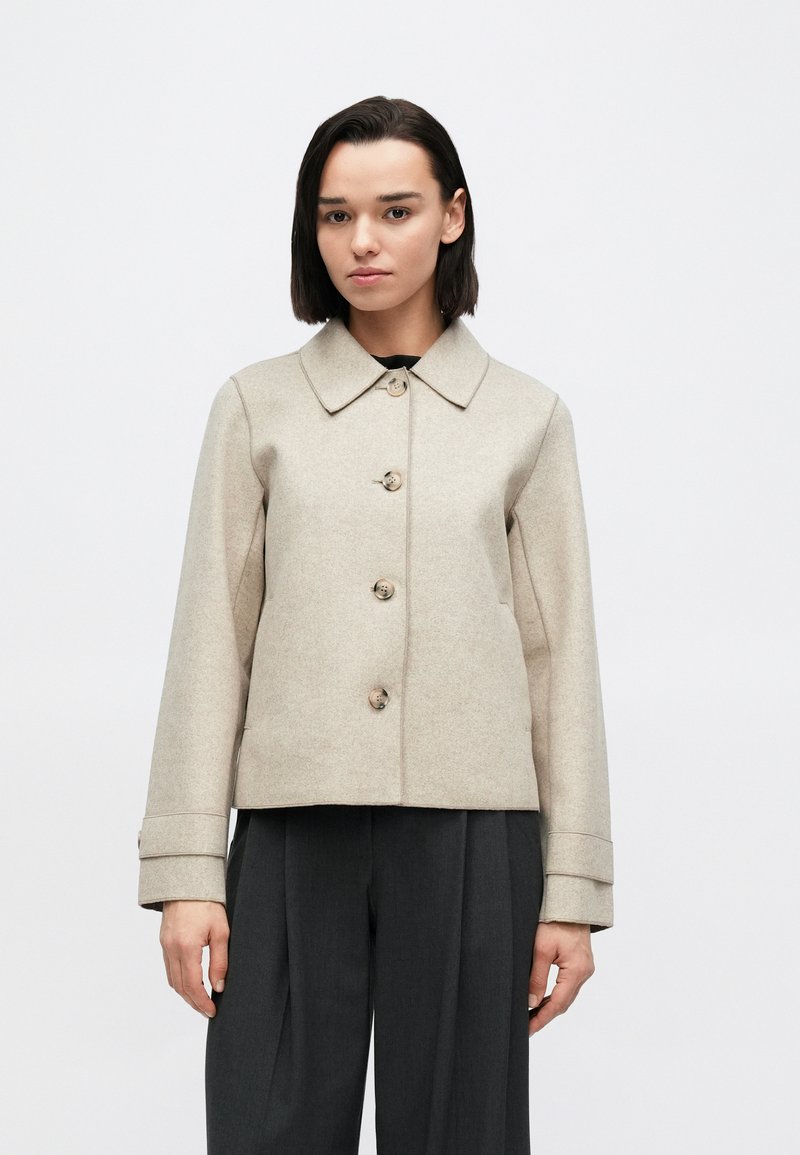 Beige jakke i ullblanding med krage, lukking med fem knapper og sidelommer. Har rett snitt og oppbrettede ermer, kombinert med mørke bukser.