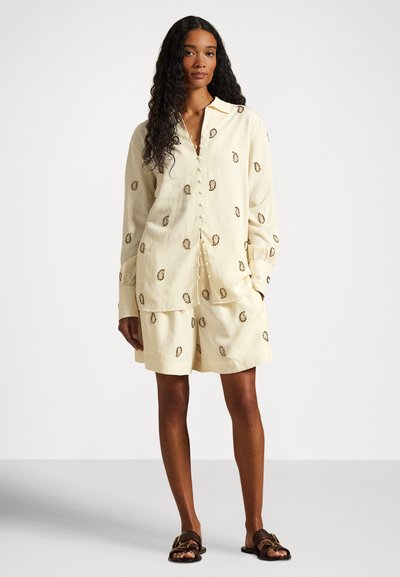 Šviesiai geltonų lino drabužių komplektas, kuriame yra marškiniai su apykakle ir mygtukais bei derančios šortai, abu dekoruoti mažais siuvinėtais raštais.