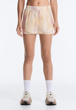 Kvinde iført lyse tie-dye løbeshorts med indbyggede beige kompressionsshorts, hvide crew-sokker og beige løbesko.