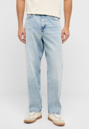 Man wearing light blue straight-leg jeans and white sneakers with beige details, standing against a plain light background.