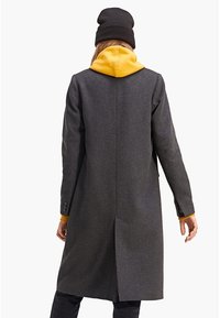 Abrigo de lana gris con una abertura en la parte trasera, que presenta una sudadera amarilla con capucha desmontable asomando. Gorro de punto negro puesto en la cabeza.