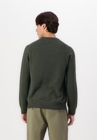 Mørkegrønn genser laget av myk stoff, med rund hals, ribbestrikkede ermer og kant. Brukt sammen med lyse khaki bukser.