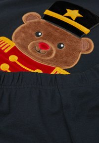 Černá tkanina s huňatým, vyšívaným medvědem v vojenském oblečení s černou čepicí a hvězdou, červeným kabátem a žlutými detaily.