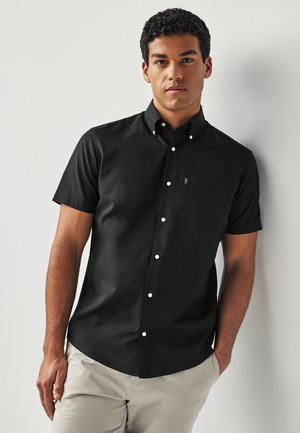 Junger Mann lehnt an einer weißen Wand, trägt ein schwarzes kurzärmeliges Hemd mit Knöpfen und hellbeige Hosen, die Hände entspannt an den Seiten.