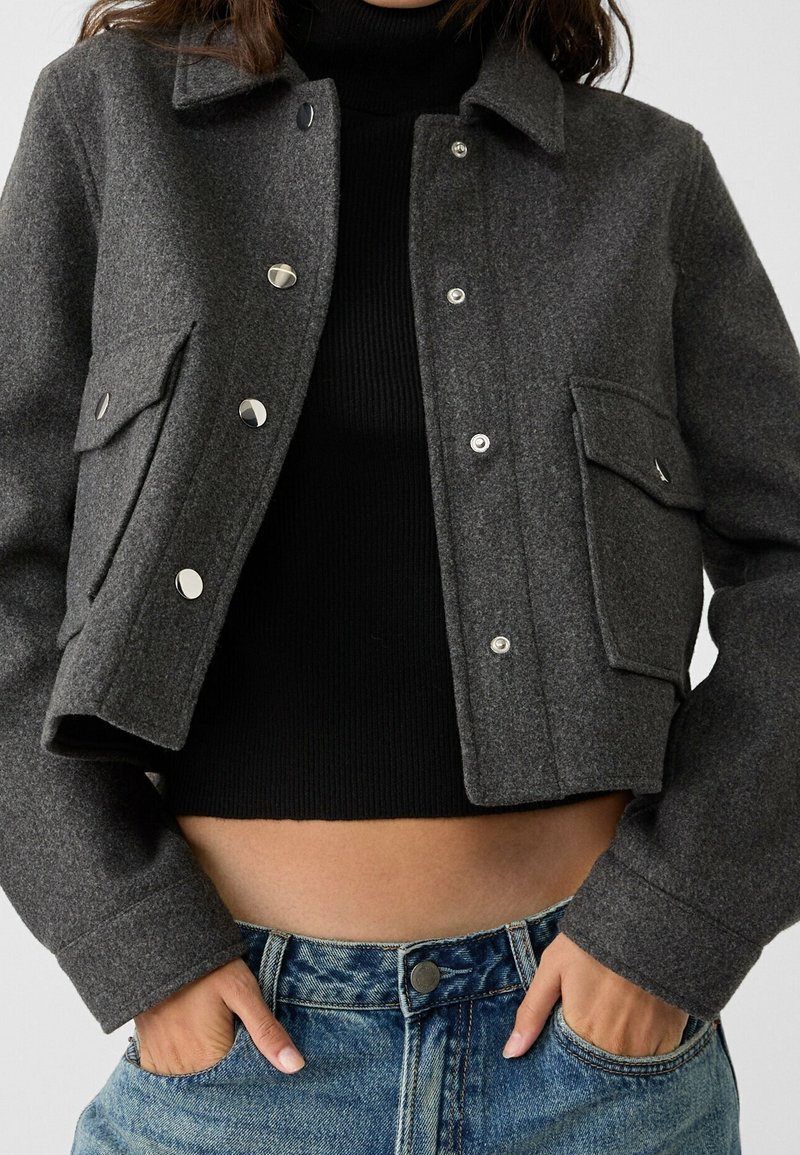 Femme portant une veste courte gris foncé boutonnée sur un pull à col roulé côtelé noir et un jean bleu, les mains dans les poches.