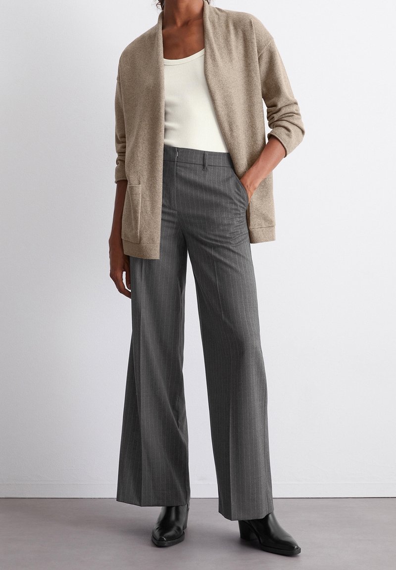 Personne portant un cardigan beige, un haut blanc, un pantalon large à fines rayures grises et des bottines noires, debout avec une main dans la poche.