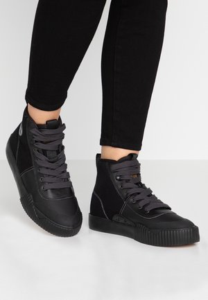 Personne portant des baskets montantes noires avec des lacets gris foncé et un pantalon noir debout sur une surface blanche.