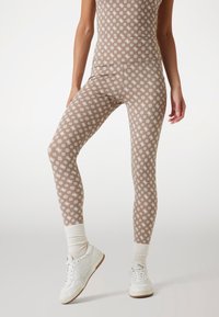 Barna mintás leggings, rugalmas anyagból készült, fehér virágmintával. Fehér tornacipővel és bordázott fehér zoknival viselve.
