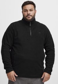 Svart fleece-pullover med halv glidelås foran, sidelommer, høy krage og teksturert stoff. Inkluderer en liten logo på brystet.