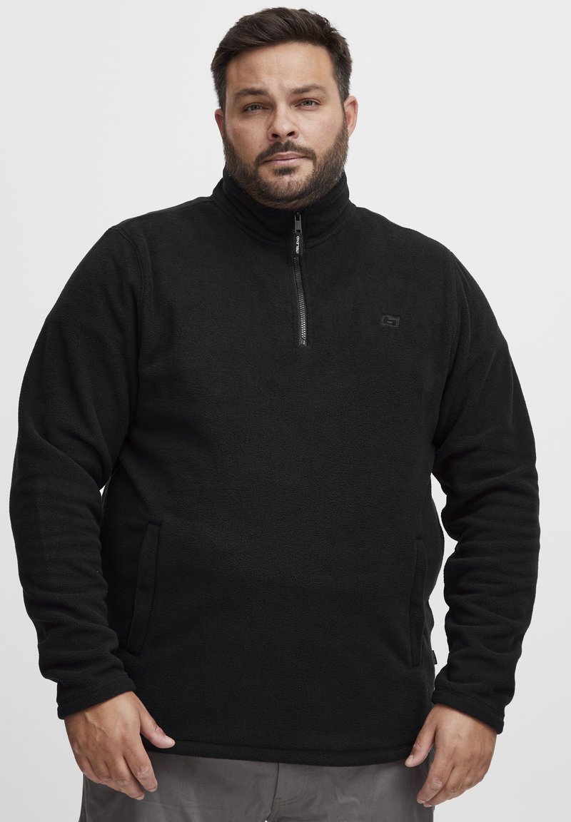 Svart fleece-pullover med halv glidelås foran, sidelommer, høy krage og teksturert stoff. Inkluderer en liten logo på brystet.