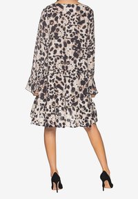 Kvinna i en knälång, långärmad klänning med ett svart och beige prickigt mönster, kombinerad med svarta högklackade skor, sedd bakifrån.