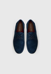 Σκούρο μπλε σουέτ παπούτσια με στρογγυλή φόρμα, που διαθέτουν σχέδιο με κορδόνια, λευκή καουτσούκ σόλα και κόκκινο εσωτερικό. Ορατό λογότυπο Tommy Hilfiger.