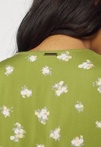 Gros plan du haut du dos d'une personne portant une chemise verte à fleurs avec une petite étiquette métallique Billabong près de l'encolure.
