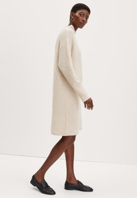 Knielanges, cremefarbenes Strickkleid mit langen Ärmeln und lässiger Passform. Texturiertes Material mit gerippten Bündchen und Saum, kombiniert mit schwarzen Slippern.