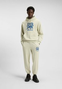 Lichtbeige hoodie en sweatpants set met blauw grafisch logo. De hoodie heeft een buidelzak; de broek heeft elastische boorden en zakken. Zwarte sneakers.