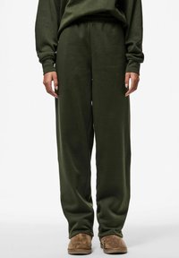 Dunkelgrüne Sweatpants aus weichem Stoff mit elastischem Bund und weiter, lässiger Passform, kombiniert mit braunen Slipschuhen.