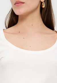 Κάτω μέρος προσώπου και λαιμός γυναίκας με πολλαπλές μικρές ελιές, φορώντας λευκό τοπ με λαιμόκοψη σε σχήμα στρογγυλεμένου V και χρυσό κρίκο στο αυτί, με μακριά καστανά μαλλιά.