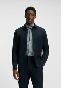 Giacca button-up blu navy sopra una camicia a scacchi bianca e nera, con taschino al petto e tessuto liscio per una vestibilità su misura.