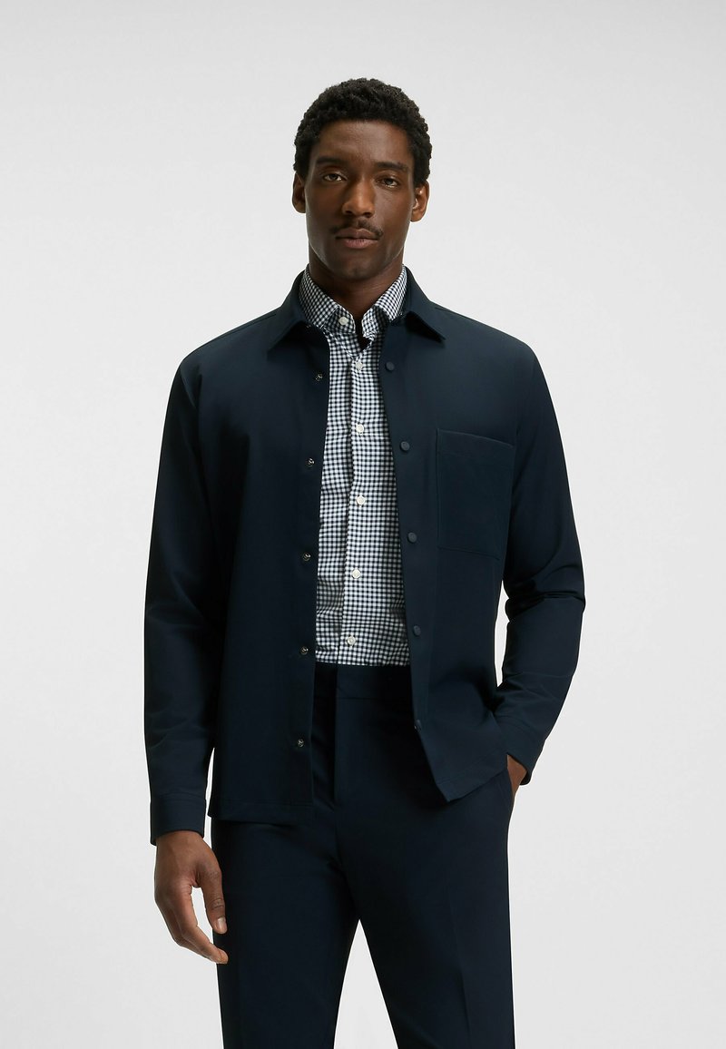 Giacca button-up blu navy sopra una camicia a scacchi bianca e nera, con taschino al petto e tessuto liscio per una vestibilità su misura.
