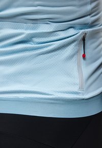 Ελαφρώς μπλε υφασμένο ύφασμα με κάθετη τσέπη φερμουάρ με μαύρο πέλμα και κόκκινη λεπτομέρεια με χάντρα, πάνω από μαύρο παντελόνι.