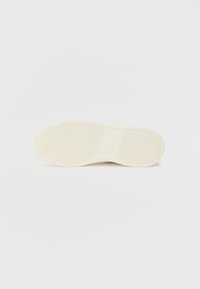 Semelle d'une chaussure, en caoutchouc crème avec une surface texturée, présentant des lignes contrastantes et une finition lisse le long des bords.