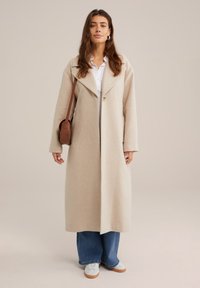 Beige ullblandad kappa med bred krage och enstaka knappstängning; kombinerad med en vit skjorta, blå vida jeans och vita sneakers.