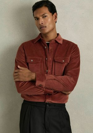 Cămașă din corduroy de culoare ruginie, cu două buzunare pe piept și manșete cu butoane, asortată cu pantaloni negri. Textură netedă și design structurat.