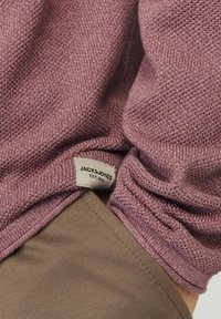 Hellvioletter Strickpullover mit strukturierter Stoffoberfläche, versehen mit einem sichtbaren weißen Etikett mit der Aufschrift "JACK & JONES EST. 1990", kombiniert mit einem beigen Kleidungsstück darunter.
