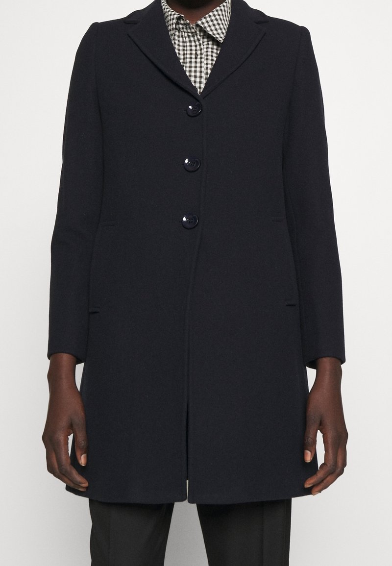 Marinblå ullkappa med en krage med snibb, fem knappar och sidofickor. Har en skräddarsydd passform och en mjuk textur. En rutig skjorta syns under.
