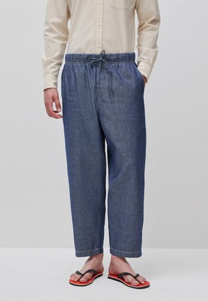Homme portant une chemise à manches longues texturée beige clair, un pantalon bleu ample à cordon et des tongs rouges à brides noires, debout sur un fond uni.
