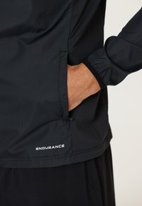 Svart vattentät jacka med strukturerad yta, elastiska ärmslut och sidficka. "ENDURANCE"-logotyp tryckt på nederkanten.