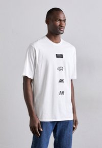 Fehér pamut t-shirt, rövid ujjú, kerek nyakkivágással. Az elülső részén fekete logók vannak, különböző méretekben és stílusokban, egymásra helyezve. A modellen kék farmernadrág van.