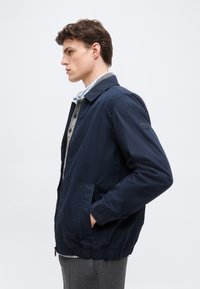 Νεαρός άνδρας με ναυτικό σακάκι, τα χέρια στις τσέπες, φορώντας κάτω από αυτό γκρι πουλόβερ με κουμπιά και ανοιχτό μπλε πουκάμισο με γιακά.