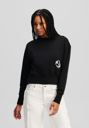 Sort cropped sweatshirt med lange ærmer og rund hals, med et hvidt logo på brystet. Matcher med hvide slidte jeans.
