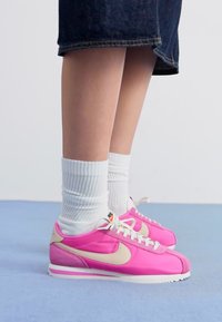 Rožiniai Nike sportbačiai su baltais raišteliais, bežiniu swoosh ir tekstūruota medžiaga. Derinami su baltais banguotais kojinėmis ir džinsine sijonu.