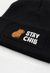 Black knit beanie with a rectangular patch featuring a brown capybara illustration and white text "STAY CHILL" in a bold font.