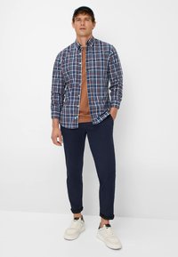 Ruitenhemde in marineblauw, rood en wit; bruine crew neck t-shirt; marineblauwe broek; witte sneakers met zwarte accenten; baseballpet.