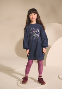 Abito felpa oversize blu navy con grafica di stella colorata e dettagli scintillanti; abbinato a leggings viola e scarpe rosse.